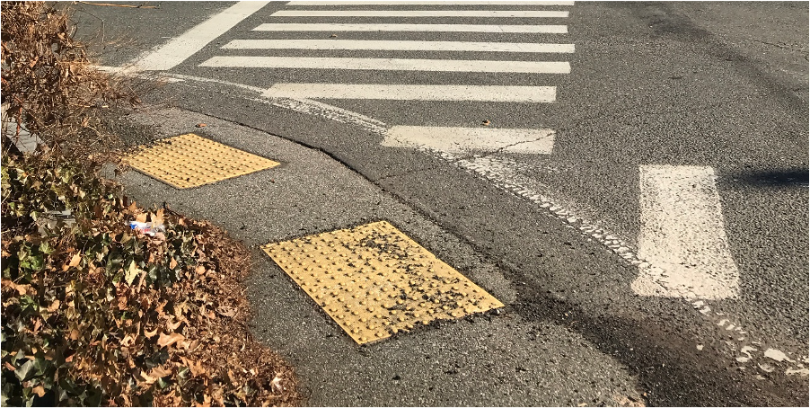Figure 9 - Northwest Intersection Pedestrian Facilities. Image shows aerial view of two crosswalks meeting on the street corner where there are two pedestrian detectable warning strips and the sidewalk is made of pavement. Figure 9 - Northwest Intersection Pedestrian Facilities. Image shows aerial view of two crosswalks meeting on the street corner where there are two pedestrian detectable warning strips and the sidewalk is made of pavement.
